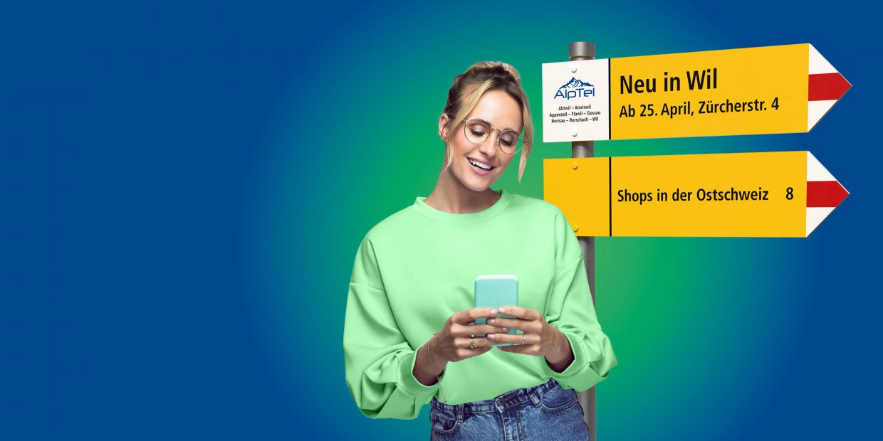 ine lächelnde Frau in grünem Pullover schaut auf ihr Smartphone. Neben ihr steht ein Wegweiser mit AlpTel-Logo und den Hinweisen „Neu in Wil – Ab 25. April, Zürcherstr. 4“ sowie „Shops in der Ostschweiz“.